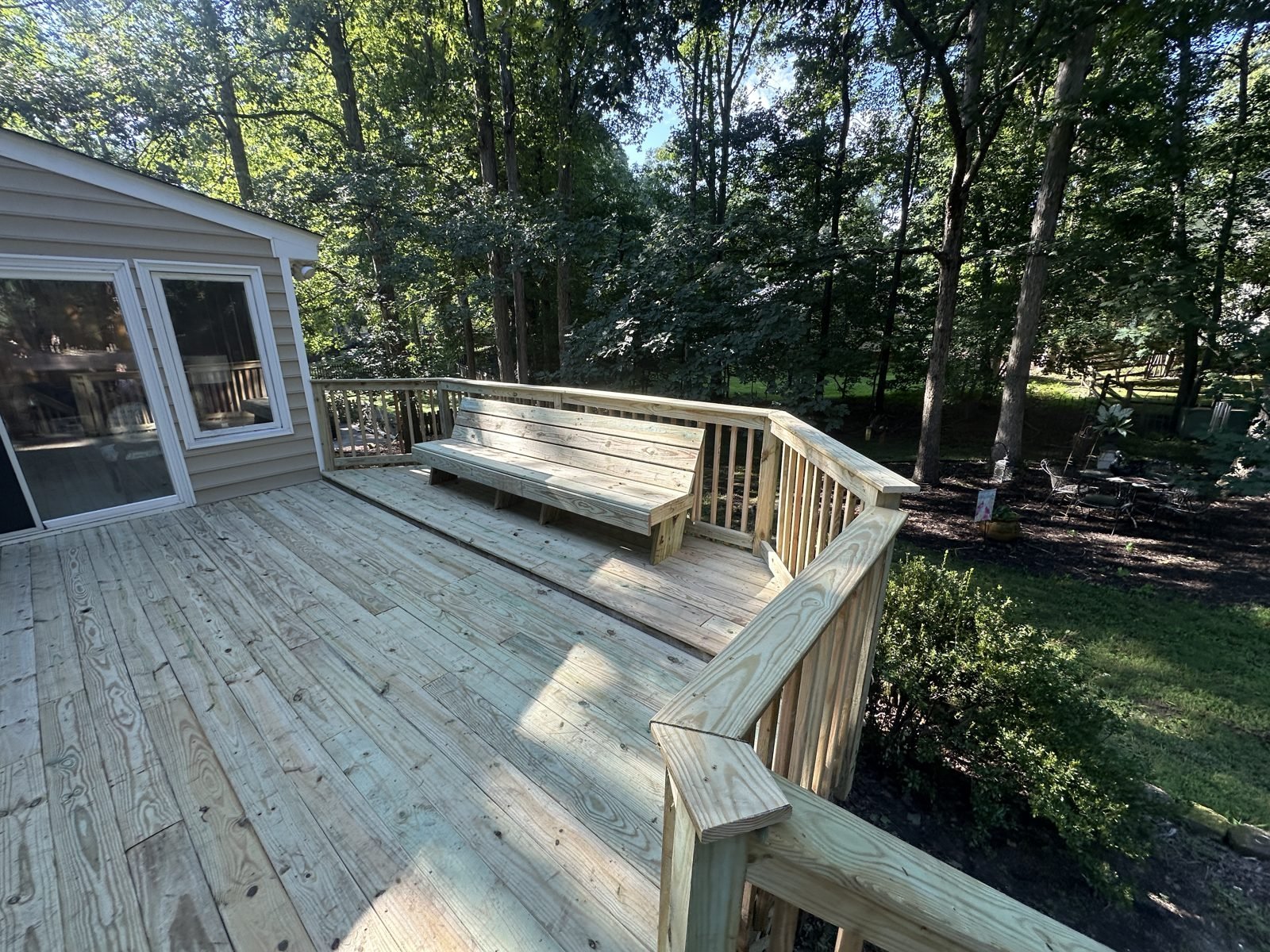 Wooden deck with railings extending from a house, overlooking forested yard with shade from tall trees