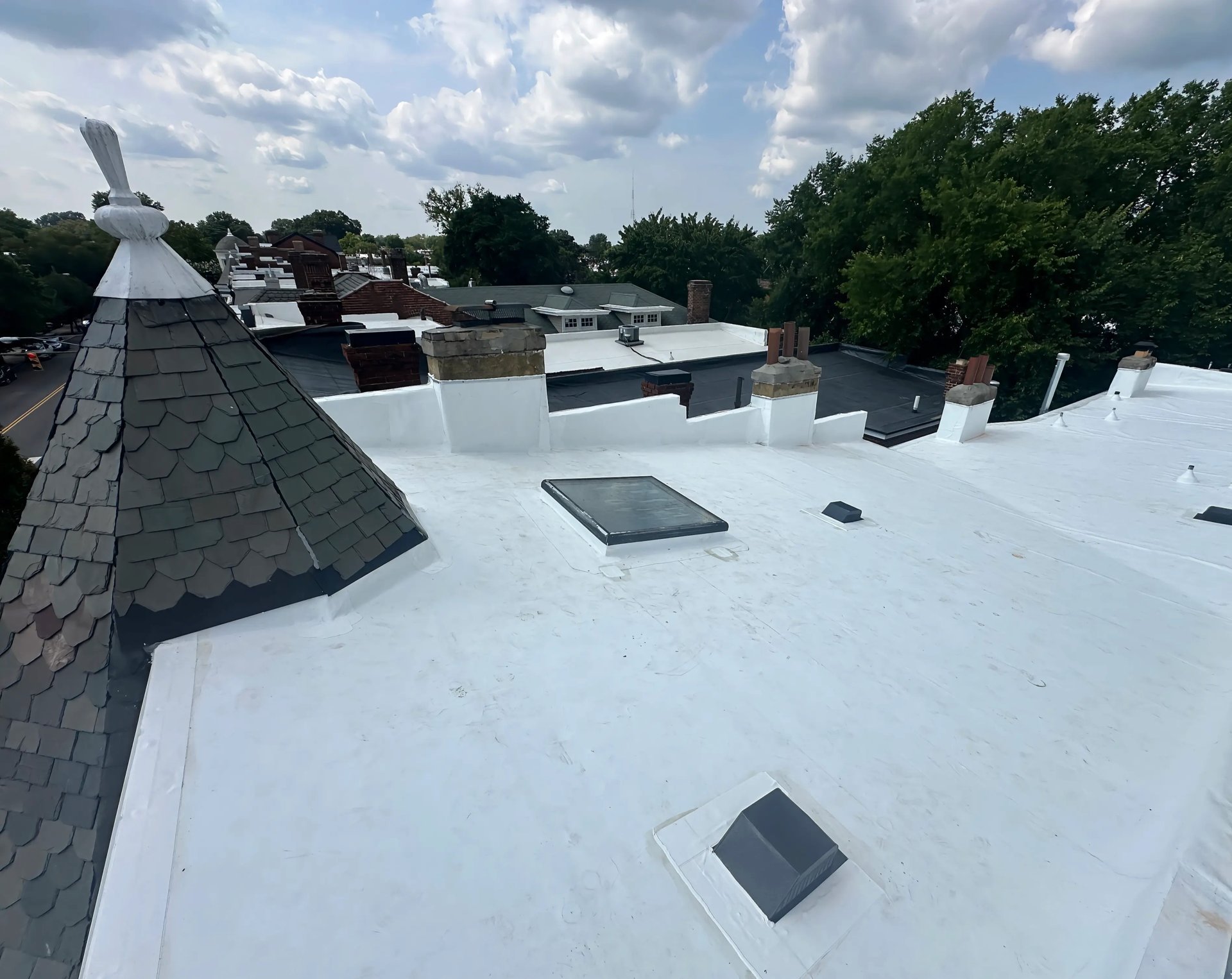 Flat white commercial rooftop with HVAC units, vents, and gray shingled peak, overlooking residential area with trees