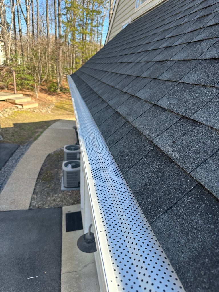 Close-up of dark gray asphalt roof shingles and white perforated gutter guard along a residential homes roofline with trees in background.