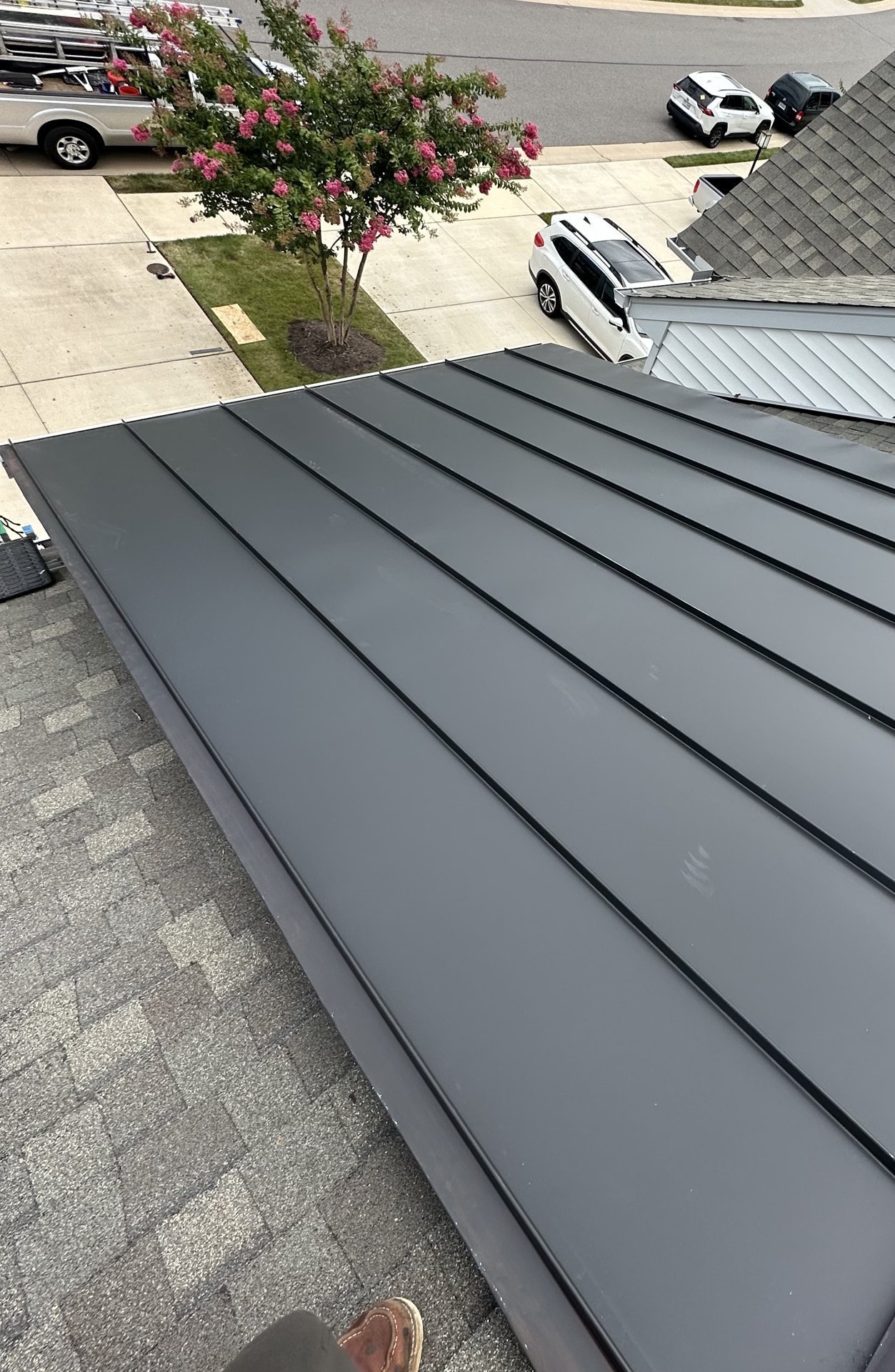 Close-up of dark gray standing seam metal roof with residential street, flowering tree, and parked cars visible below