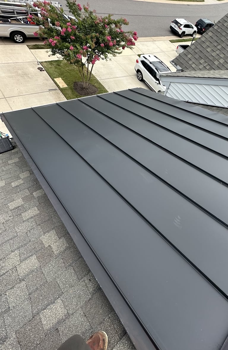Close-up of dark gray standing seam metal roof with residential street, flowering tree, and parked cars visible below