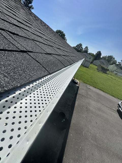 Perforated metal gutter system installed along a residential roof with asphalt shingles, green lawn and trees visible in background