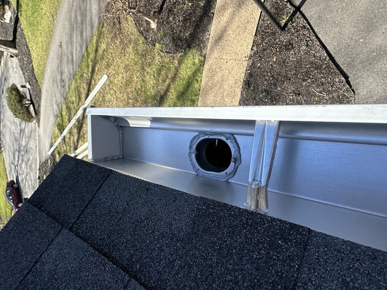 Close-up of a residential gutter with drainage pipe and black shingles on roof edge, viewed from above