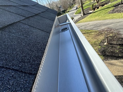 Close-up of a residential gutter with downspout along a shingled roof edge, showing landscaped yard in background