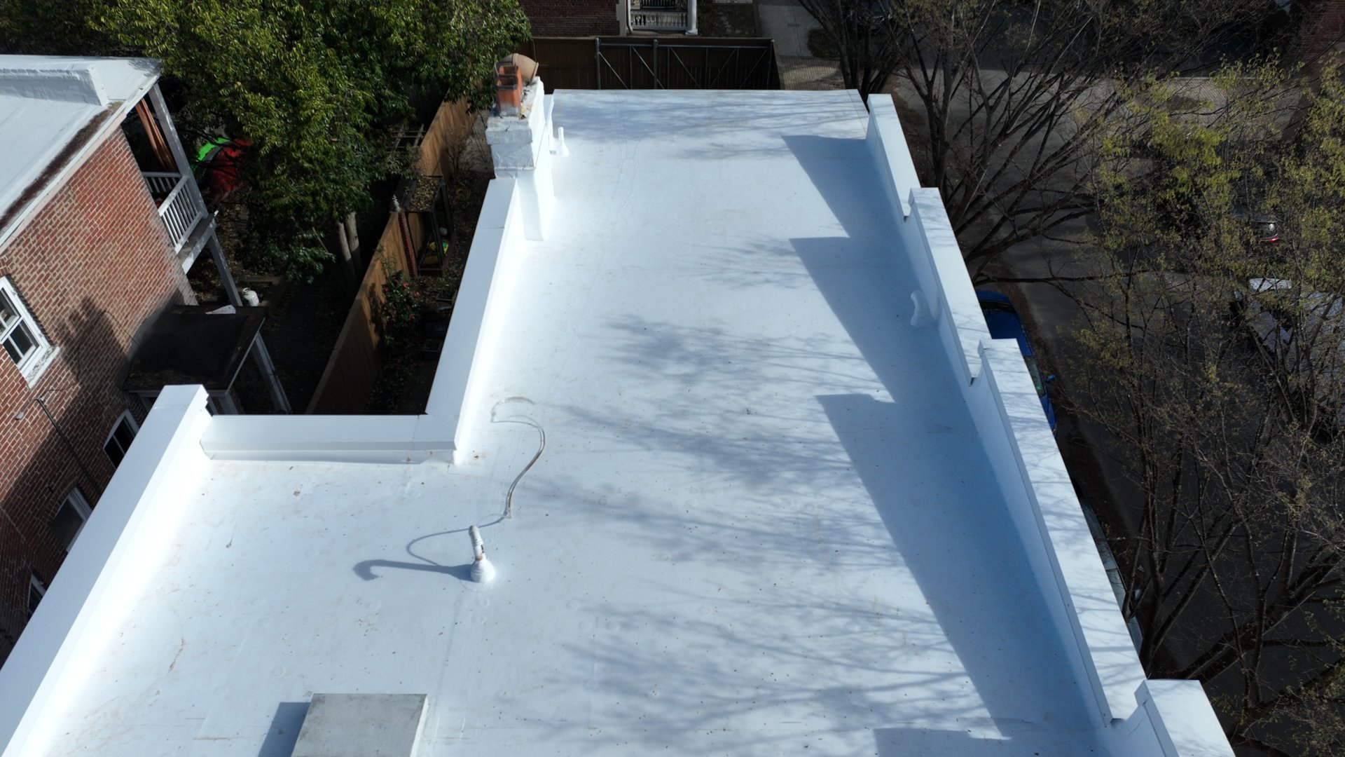 Aerial view of a flat white roof with shadows, surrounded by brick building, trees, and a parked car