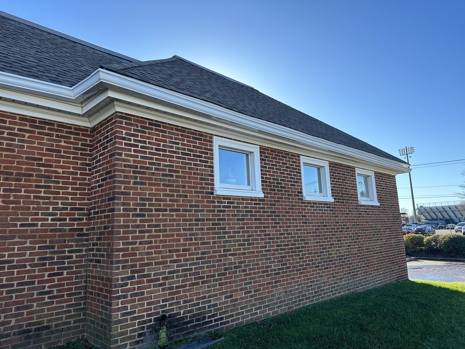 A modern brick residential building with dark roof, three white windows, green lawn, and clear blue sky.
